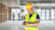 construction worker in the foreground wearing a yellow hard hat and a high-visibility safety vest looks down at his smartphone.