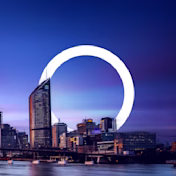 A cityscape at sunset with a river, a bridge, and buildings, one of which is encircled by a glowing ring.