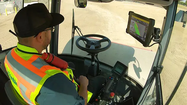 A view from the back of a vehicle cab, showing a person wearing a black hat and a bright orange and yellow safety vest, seated in the driver's seat.