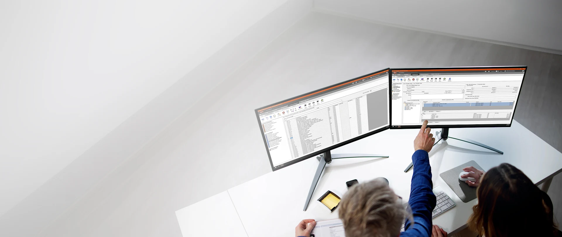 Two people use Trimble Viewpoint Estimating software on dual monitors to analyze construction project data at a white desk.