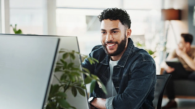 A man with a beard smiles while looking at a desktop computer in an office setting.