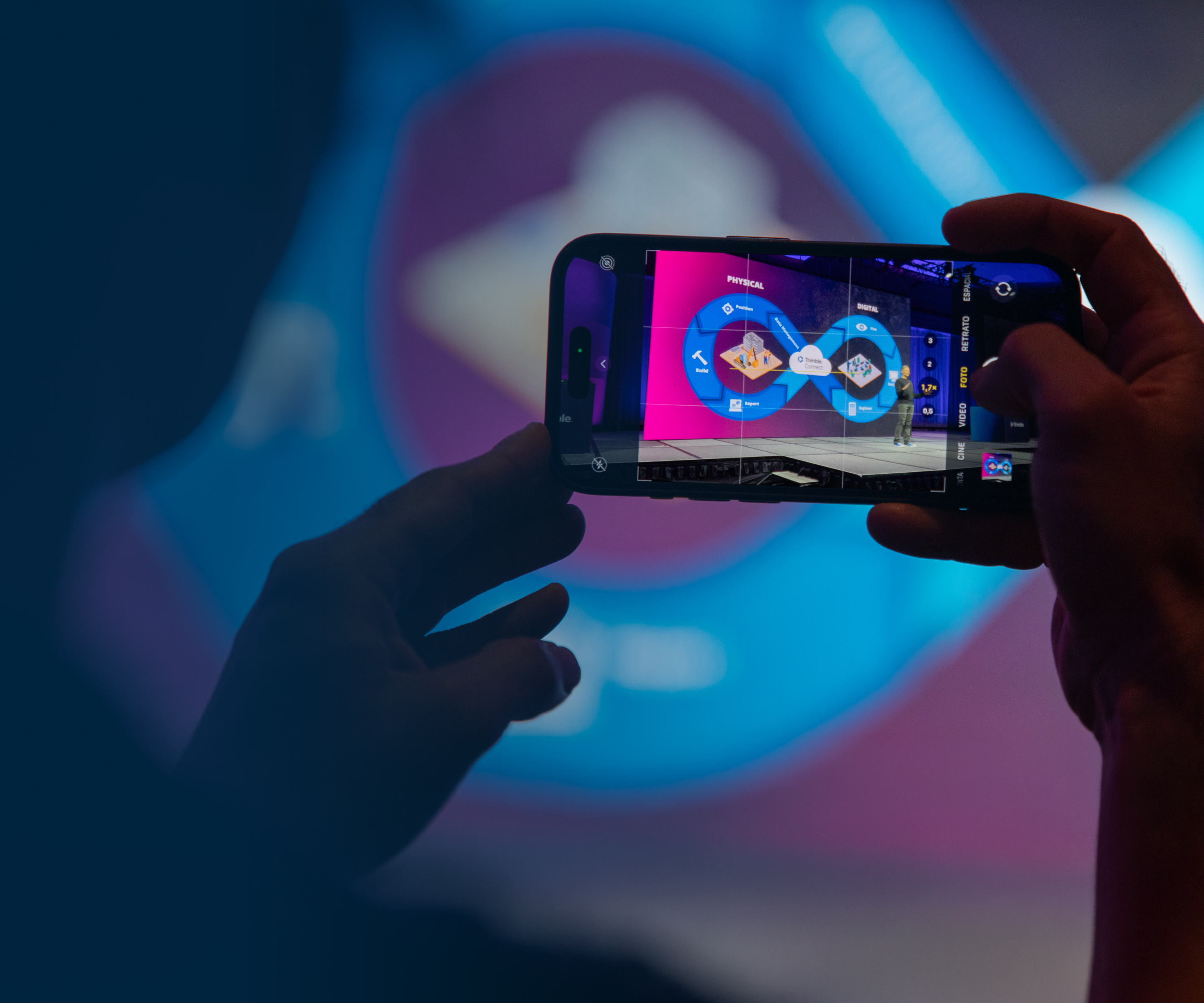A person uses a smartphone to photograph a speaker and a digital presentation on stage at the Trimble Dimensions conference.