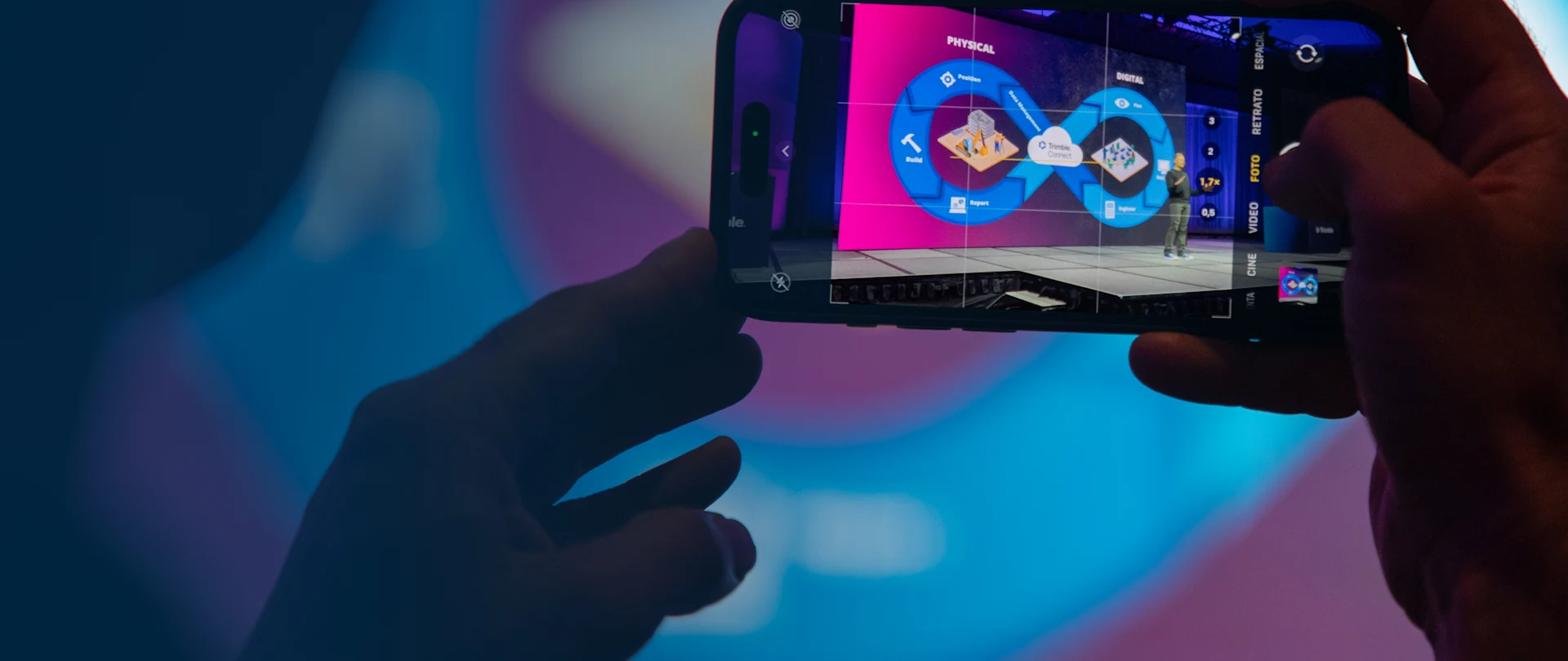 A person uses a smartphone to photograph a speaker and a digital presentation on stage at the Trimble Dimensions conference.