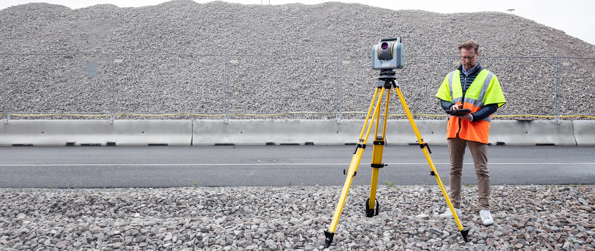A surveyor in a high-vis vest operates a Trimble SX10 scanning total station on a yellow tripod near a large gravel heap.