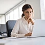 Pensive woman in a white blouse working on a laptop in a bright office, resting her chin on her hand.