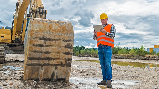 A construction worker in a safety vest uses a Trimble tablet near an excavator bucket at a job site to manage machine guidance and site data.
