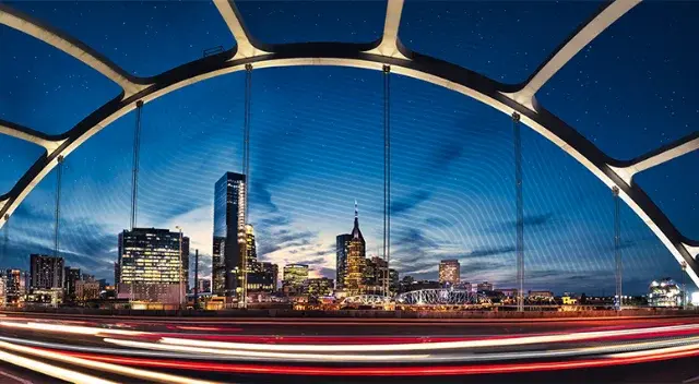 Nashville skyline through the John Seigenthaler Pedestrian Bridge's concrete arches at dusk. Active traffic lights streak foreground. AT&T "Batman Building" symbolizes urban construction.