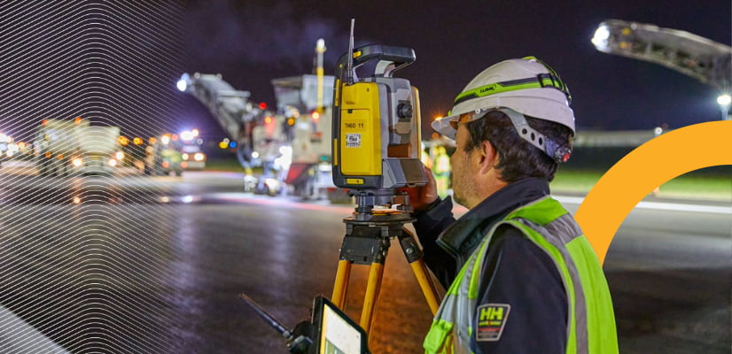 A surveyor in safety gear uses a total station at a construction site at night with heavy machinery.
