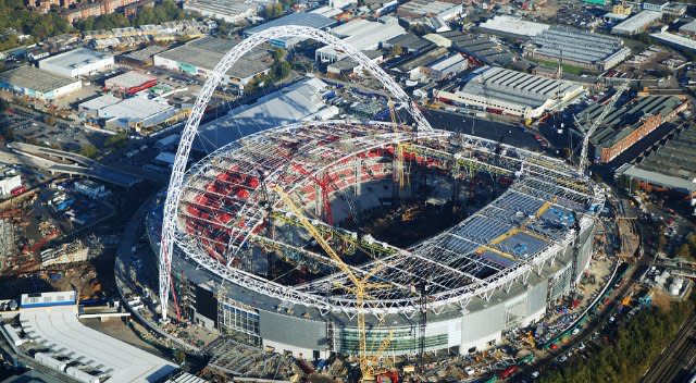 Vue aérienne du stade de Wembley à Londres en cours de construction. L’imposante arche d'acier de 133 m de hauteur soutient le toit partiellement installé. Les grues et la charpente métallique déployée reflètent l'avancement du projet AECO.