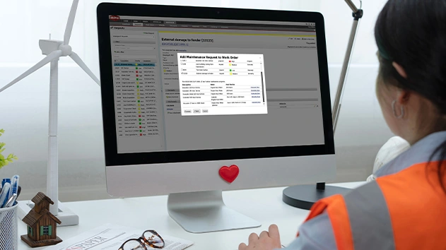 A woman in a safety vest uses Trimble TMT maintenance software at a desk with model wind turbines and a heart-stickered monitor.