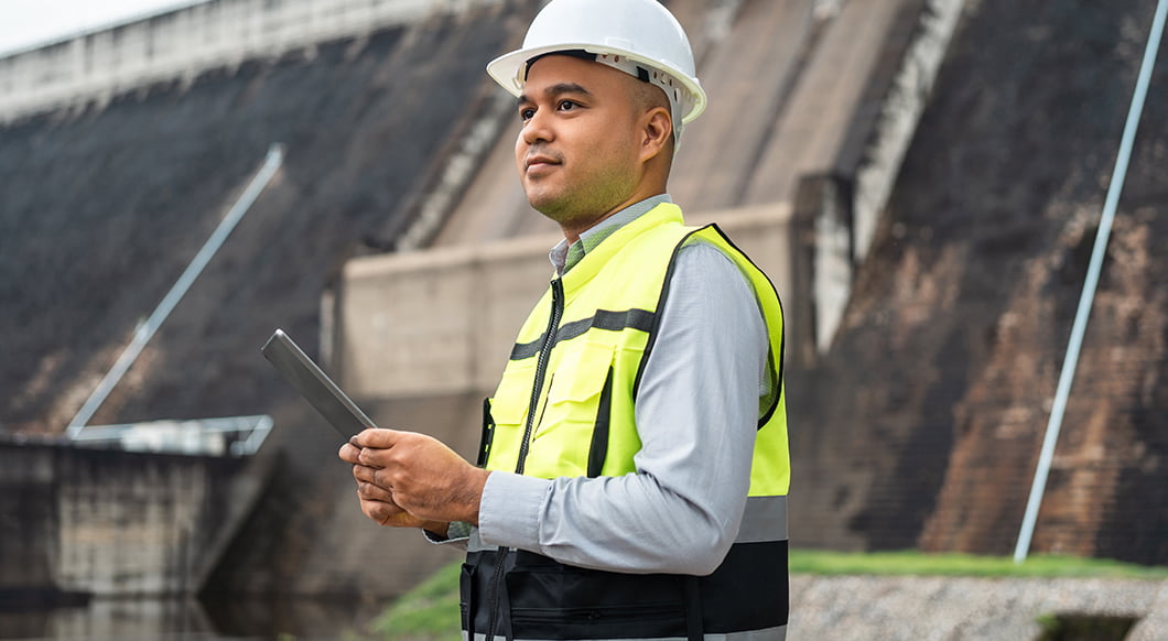 A male engineer in a white hard hat and yellow safety vest holds a tablet in front of a large concrete dam.