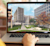 A person uses a laptop to view an architectural rendering of a modern building complex with a grassy slope, autumn trees, and stylized figures.