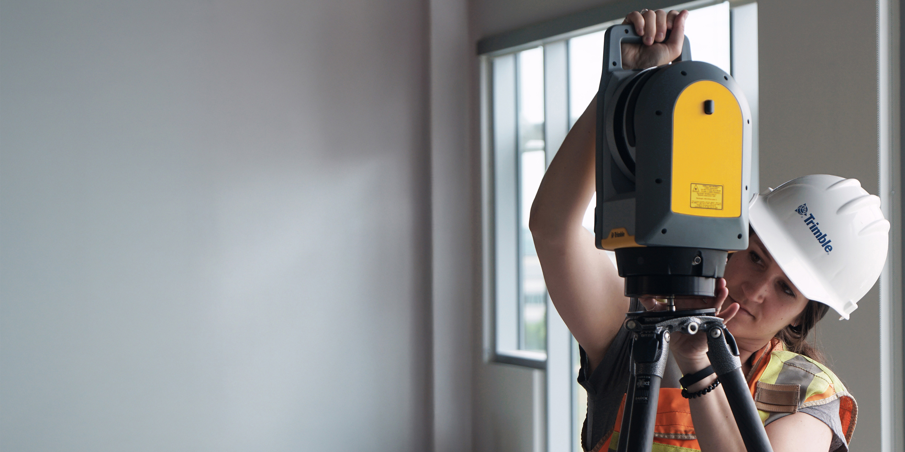 A woman in a hard hat and safety vest sets up a Trimble laser scanner on a tripod inside a building under construction.