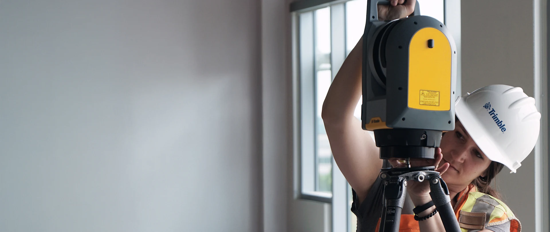 A woman in a hard hat and safety vest sets up a Trimble laser scanner on a tripod inside a building under construction.