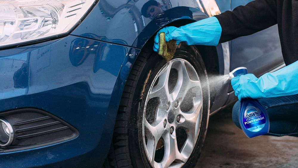 A person wearing blue rubber gloves spraying Dawn Powerwash dish spray onto the rim while wiping it with a green sponge