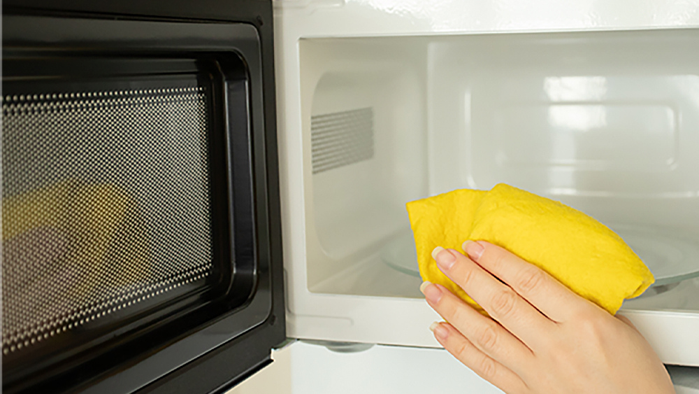 Hand wiping the inside of a microwave with a yellow cleaning cloth.