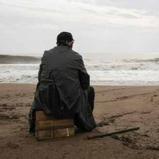 Une personne âgée est assise sur une pile de bois et regarde l’océan d’un air pensif