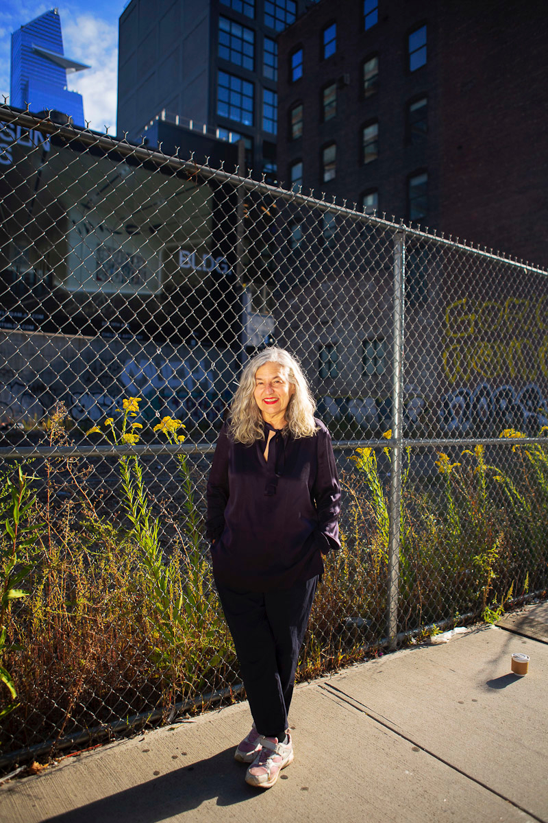A woman stands in front of a fence with yellow flowers on the sidewalk