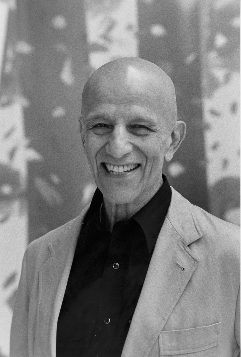Black and white photo of a man smiling, wearing a jacket and button down shirt in front of a painting