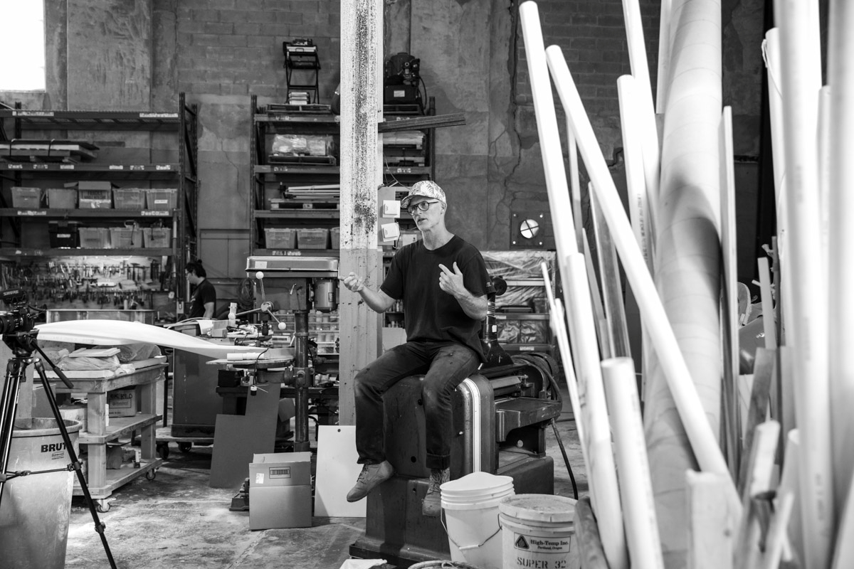 Black and White photo of a seated man wearing a hat and glasses in midst of speaking, surrounded by artist's studio setting