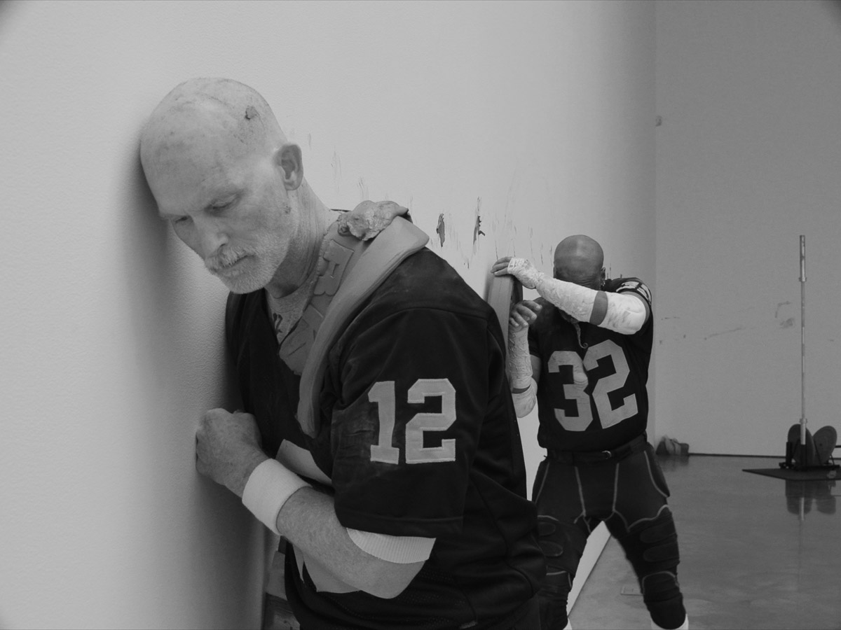 Black and white image of two men wearing black football jerseys performing in a white room with a concrete floor