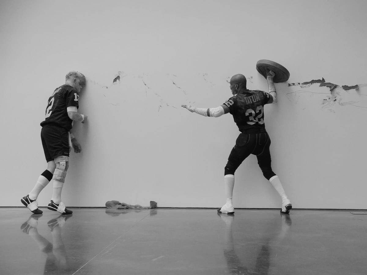 Black and white image of two men wearing football jerseys performing choregraphy in a white room with a concrete floor