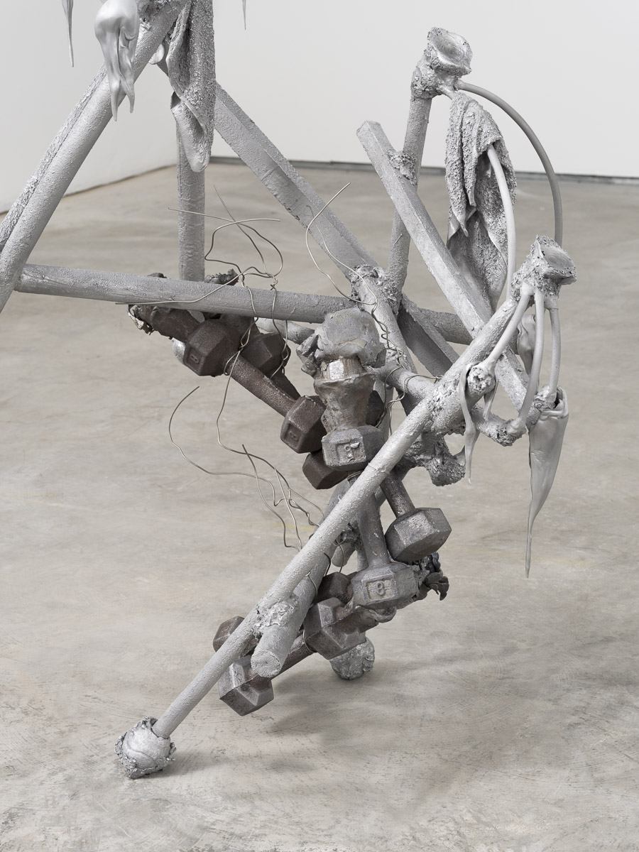 Detail view of an aluminum and lead sculpture in a white room on a concrete floor.
