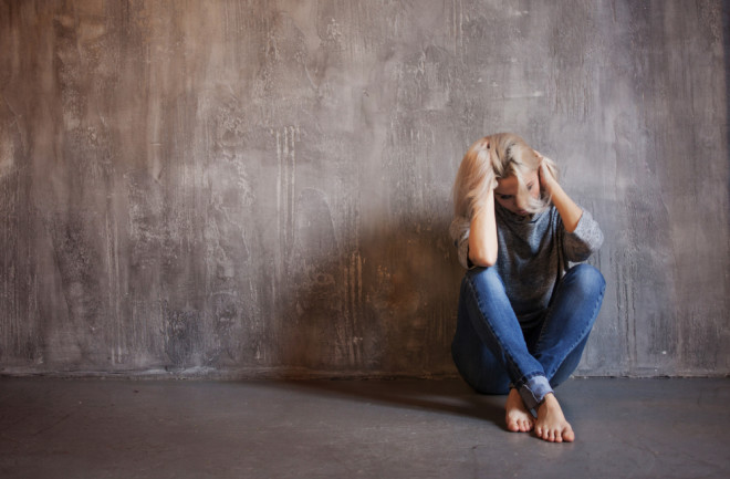 Sad woman sits on the floor. Depression and chronic fatigue. Young beautiful blonde in a gray sweater and jeans, gray textured background