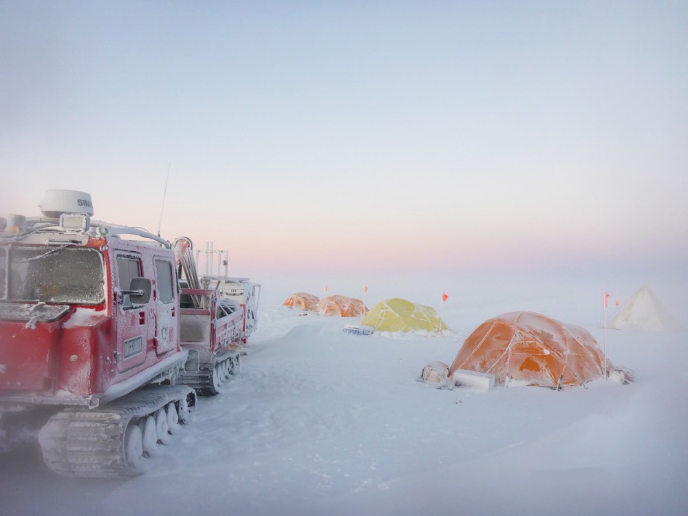 Left, a red Antarctic overland vehicle sits, snow-splattered, on the ice. Right, orange and yellow tents are set up.