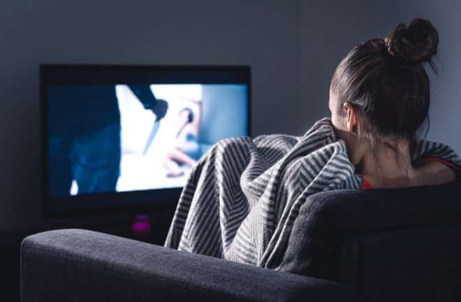 Scary horror movie on tv. Scared woman watching stream service hiding under blanket on couch at night. Sleepless person streaming series or film on television. Alone in dark and afraid of thriller.