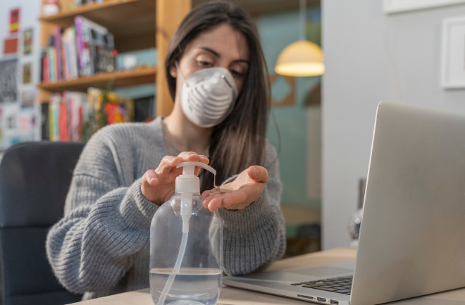 Woman at desk wearing mask and using hand sanitizer - Shutterstock