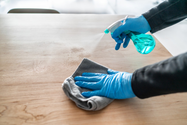 Two hands in latex gloves clean a tabletop with a spray bottle and rag - Shutterstock