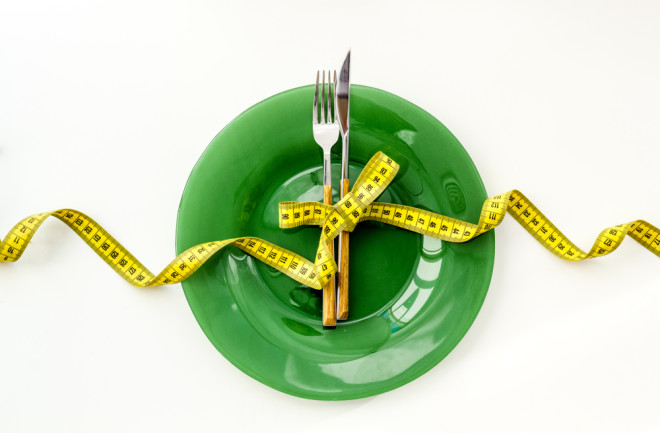 Diet Weight Loss Concept, Knife and Fork tied in a bow with measuring tape, on a green plate - Shutterstock