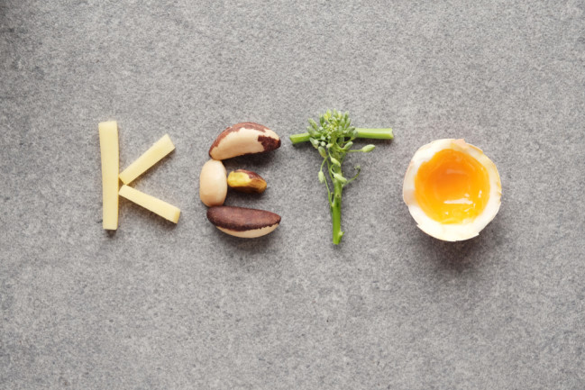 The word "KETO" spelled with food items on a grey background: "K" with fries, "E" with Brazil nuts, "T" with broccoli, and "O" with a half-boiled egg