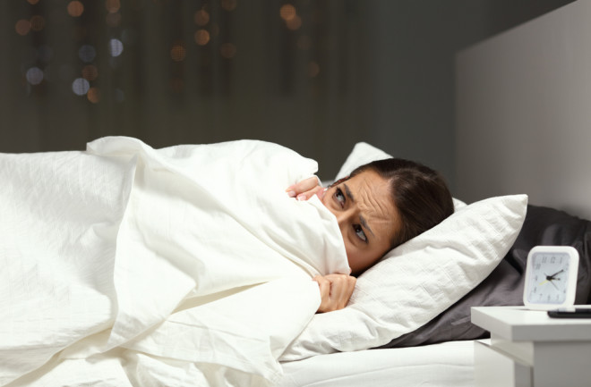 Woman in bed, scared, hiding under covers - Shutterstock