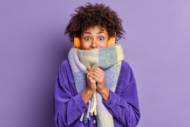 Black woman feels very cold during freezing weather wears purple jacket and warm scarf around neck wals on street during winter listens music via wireless headphones keeps hands together