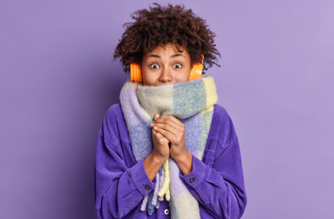 Black woman feels very cold during freezing weather wears purple jacket and warm scarf around neck wals on street during winter listens music via wireless headphones keeps hands together