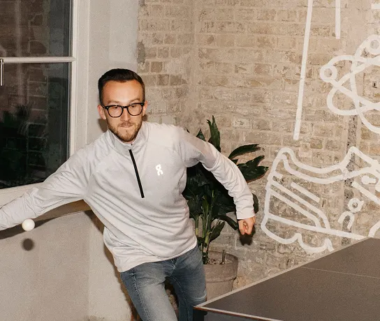 A man wearing glasses and a light pullover reaches to hit a ping-pong ball at an indoor table in front of a brick wall with a white mural.