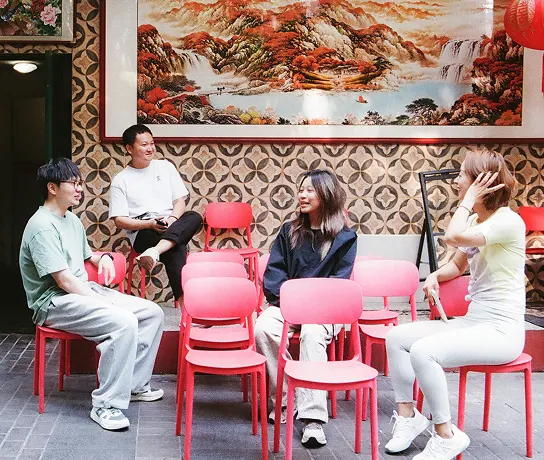 Four people sit and talk on red chairs in a room with patterned wallpaper and a large landscape painting mounted on the wall.