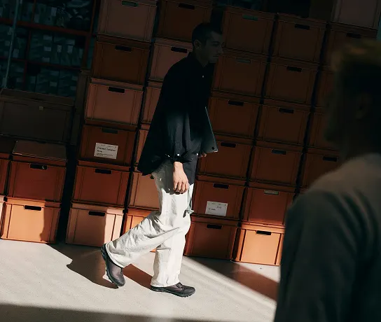 A person wearing a dark jacket and light pants walks past a wall of stacked orange storage boxes while another person is blurred in the foreground.