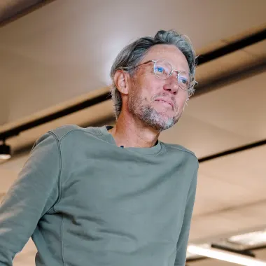 Older man with gray hair and glasses wearing a green sweatshirt, looking slightly upward in a modern indoor space with ceiling lights and panels.