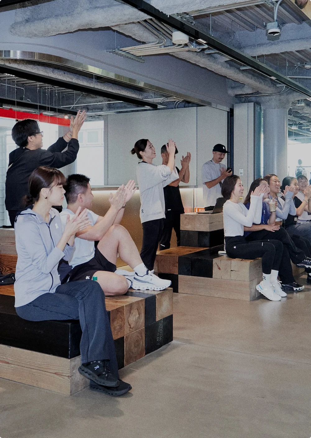 A group of people seated and standing on wooden block benches inside a modern space, clapping and smiling.
