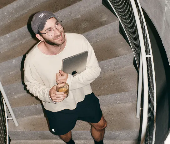 A man wearing a baseball cap, glasses, a light sweater and black shorts stands on a concrete stairwell holding a tablet under his arm and a glass while smiling and looking up.