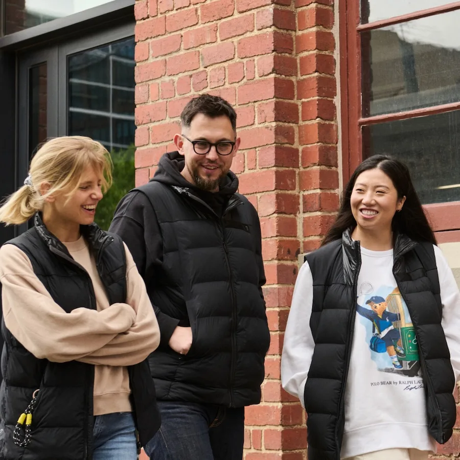 Three people wearing black puffer vests laugh and walk together outside a red brick building.