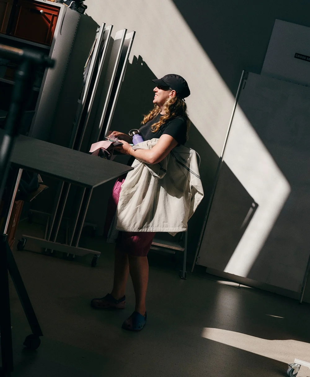 A woman wearing a black cap and dark shirt stands indoors in a diagonal patch of sunlight, holding a light jacket, a purple water bottle, and some papers.