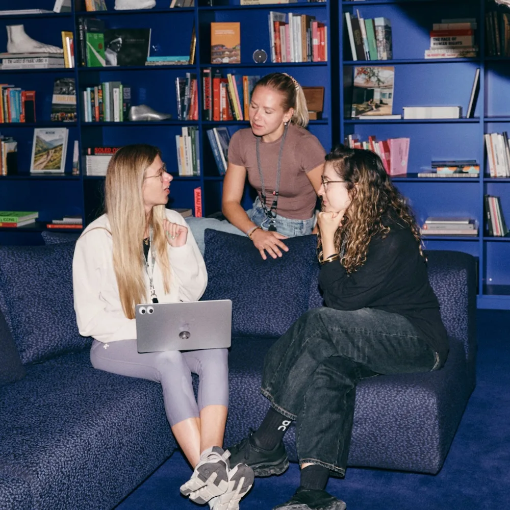 Three women chat on a dark blue couch in front of a bookshelf, one holding a laptop while another leans in.