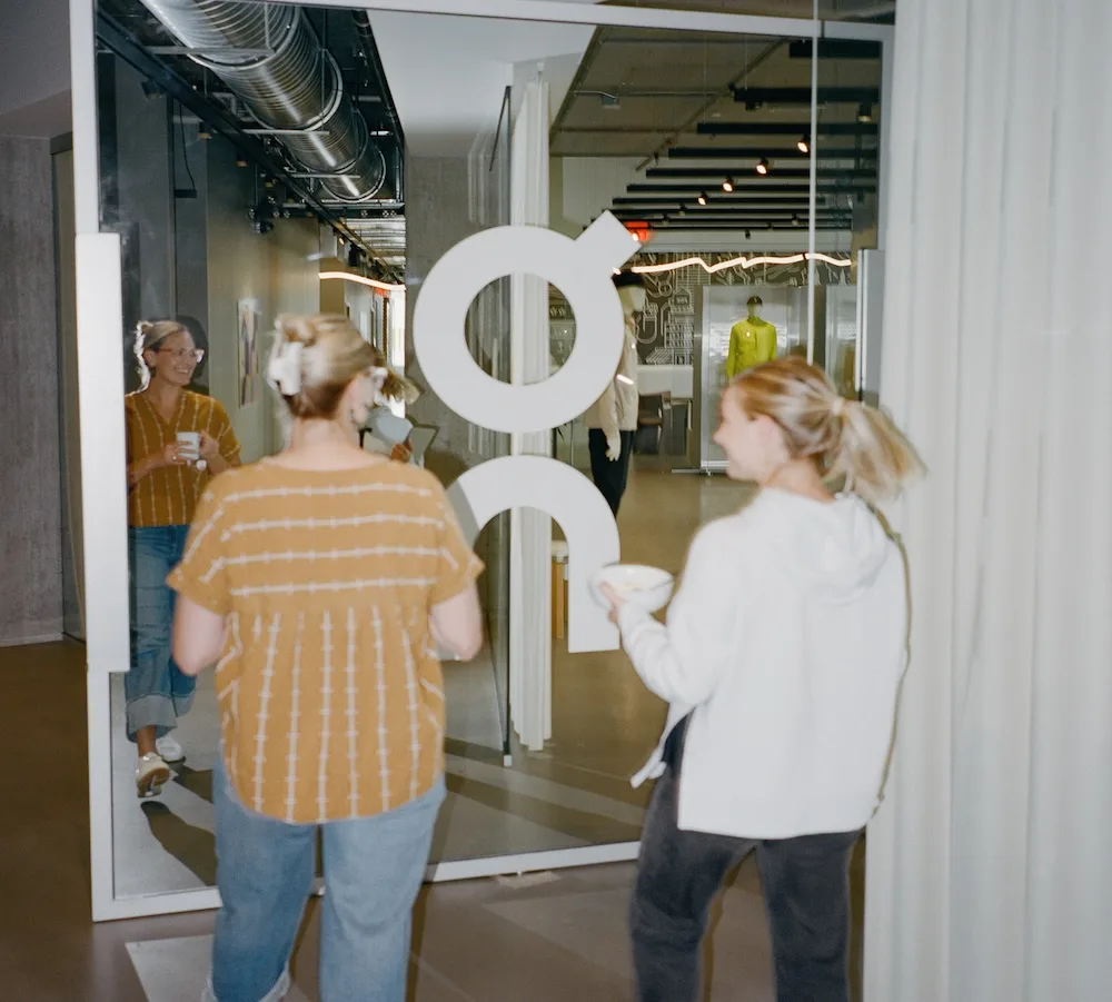 Two people enter through a glass door bearing a large white circular logo, one holding a bowl, a mirrored reflection of another person is visible, and a mannequin wearing bright yellow stands in the background.