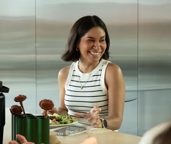 Three people sitting on a long curved sofa at small round desks. A smiling woman in a sleeveless white and navy striped top sits at a table with a salad and gestures with her hand.