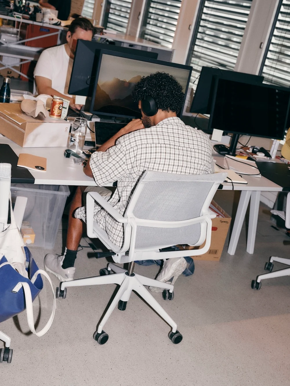 Person wearing headphones works at a computer in a busy office with desks, packaging boxes, and supplies.