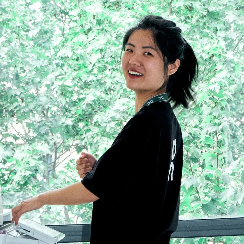 Woman in a black shirt turns to smile at the camera while using a printer beside a window filled with leafy trees.